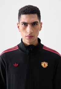 Black zip-up jacket with red accents and stripes, featuring embroidered Adidas logo and Manchester United crest on the chest.