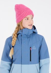 Giacca invernale blu e azzurro con colletto alto e tasca con zip, indossata con un berretto di lana rosa acceso. Materiali testurizzati visibili.