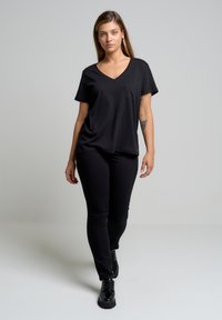 Crna majica kratkih rukava s V-izrezom i džepom, u kombinaciji s crnim uskim hlačama i crnim čizmama na vezanje, na svijetloj pozadini.