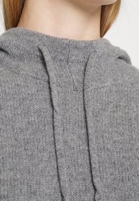 Szary sweter z dzianiny z kapturem, wykonany z fakturowanej tkaniny, wyposażony w regulowany sznurek. Widok zbliżenia na obszar szyi i ramion.