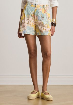 Kobieta w szortach w kwiatowe wzory w pastelowych kolorach, białej koszuli, złotych i bursztynowych bransoletkach oraz żółtych espadrylach.