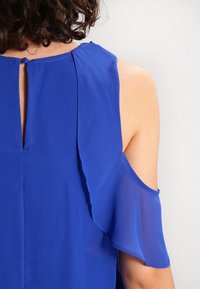 Blusa azul con cierre en la parte posterior tipo ojal, presenta mangas cortas y transparentes, y un diseño con volantes en los hombros. Textura de tela suave.