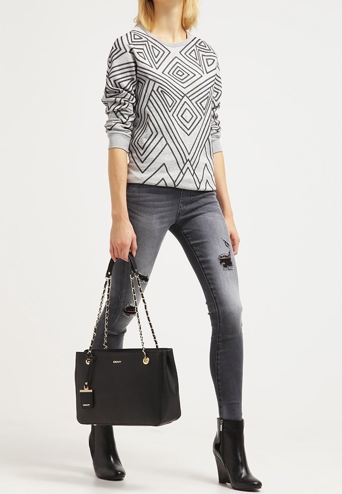Personne portant un pull gris à motifs, un jean skinny gris déchiré, des bottines noires et tenant un sac à main noir avec des anses en chaîne.