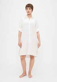 Vestito camicia bianco con maniche corte arrotolate, tessuto morbido, vestibilità ampia e spacco laterale per facilitare i movimenti.