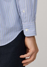 Mano de un hombre descansando a un lado, vistiendo una camisa de botones a rayas azules y blancas y pantalones oscuros con textura, contra un fondo liso.