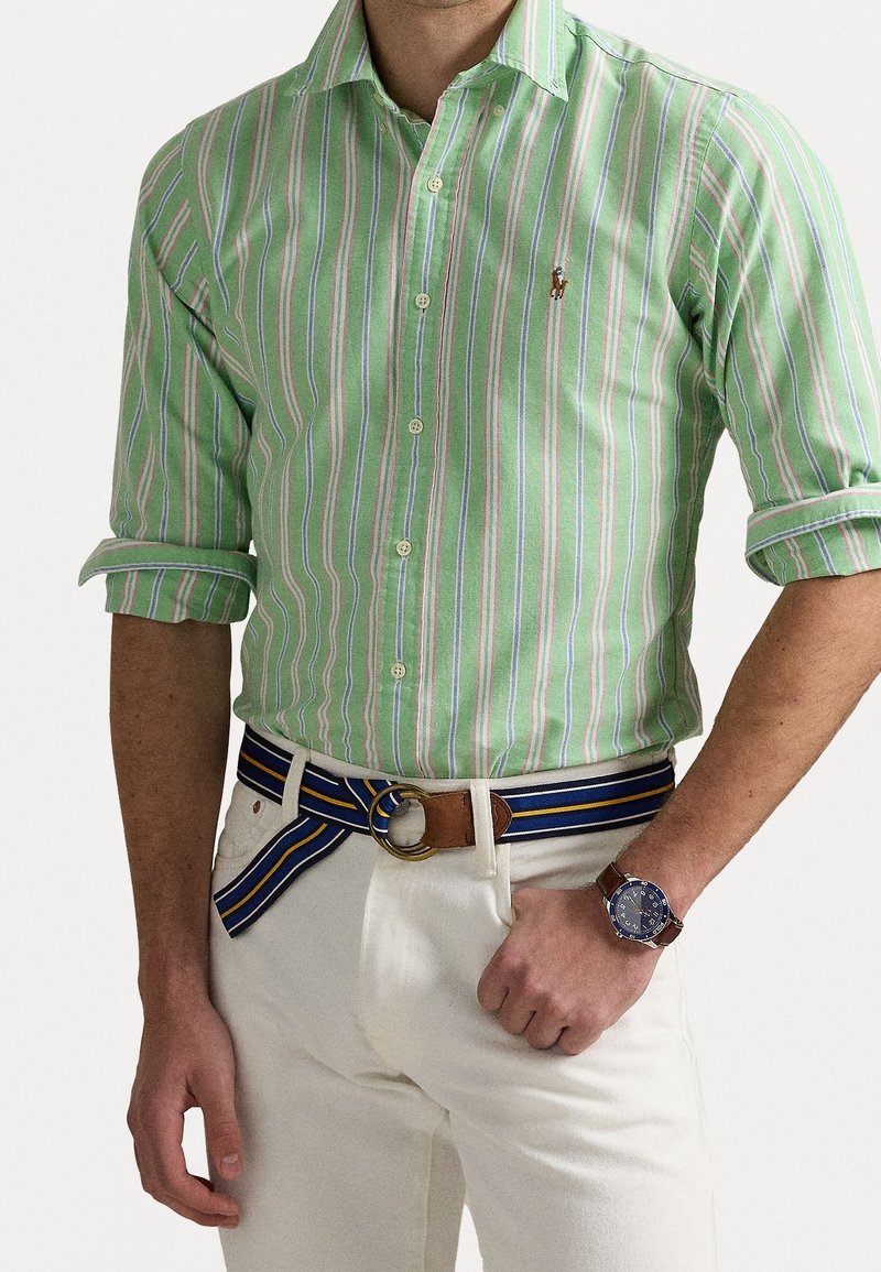 Camisa a rayas verdes con líneas blancas, rosas y azules, diseño ajustado, cuello abotonado. Combinada con pantalones beige y un cinturón de rayas múltiples.