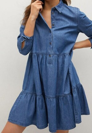 Žena v modrých džínových šatech na knoflíky s vrstvenou sukní a dlouhými rukávy vyhrnutými k loktům, jedna ruka u límce.