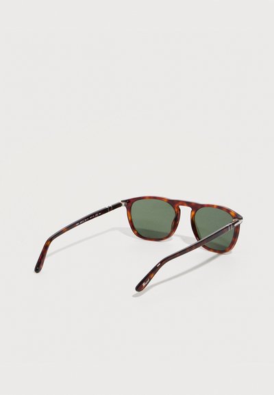 Γυαλιά ηλίου με σκελετό χελώνας και σκοτεινούς πράσινους ορθογώνιους φακούς, ανοιχτά πάνω σε ανοιχτό γκρι επιφάνεια.