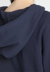 Granatowa bluza z kapturem o miękkiej, teksturowanej tkaninie, w luźnym kroju i prostym designie, podkreślająca konstrukcję kaptura.