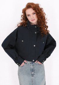 Marineblauwe cropped jack met een hoge kraag, grote voorzakken en drukknoopsluiting. De stof lijkt soepel en lichtgewicht. Gecombineerd met een denim rok.