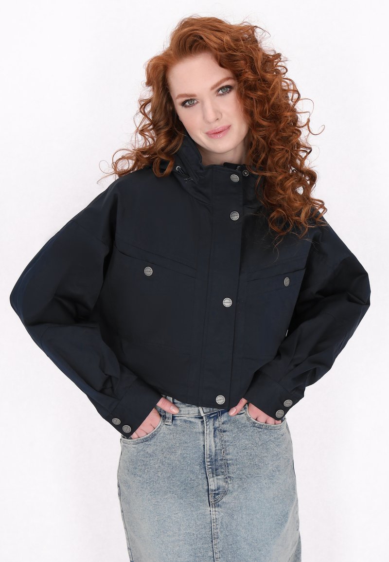 Marineblauwe cropped jack met een hoge kraag, grote voorzakken en drukknoopsluiting. De stof lijkt soepel en lichtgewicht. Gecombineerd met een denim rok.