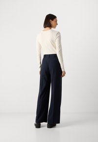 Maglietta a maniche lunghe color crema con scollatura rotonda abbinata a pantaloni a vita alta a righe blu marino. Modella in piedi di spalle, indossa scarpe con piattaforma nere.