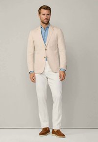 Mężczyzna w beżowej marynarce, niebieskiej koszuli, białych spodniach i brązowych zamszowych mokasynach stojący na tle jasnoszarym.