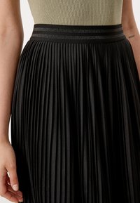 Svart plisserad kjol med en slät yta, med en kontrasterande midja med två metalliska ränder. Närbild visar detaljerad plissering.