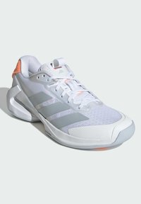 Bílá sportovní obuv se šedými třemi pruhy, svršek z prodyšné síťoviny, gumová podrážka a oranžové detaily na patě a jazyku. Má zavazovací design.