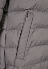 Grå vadderad jacka med ett vertikalt dragkedjefack delvis öppet, vilket visar dragkedjens dragare och sömnadsdetaljer.