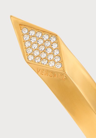 Pointe d’accessoire Versace ton or avec un groupe en forme de diamant de cristaux clairs et un logo Versace gravé sur une surface dorée lisse.