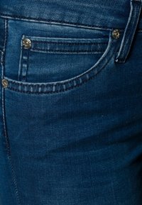 Jean en denim bleu foncé avec une texture lisse, doté d'une poche avant, de rivets argentés et de détails cousus le long des bords.