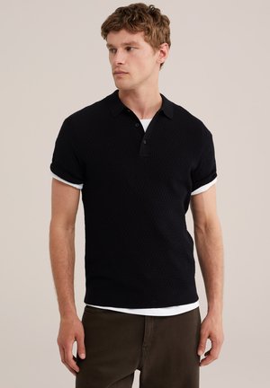 Jeune homme aux cheveux courts et bouclés portant un polo noir texturé à manches courtes par-dessus un t-shirt blanc, assorti à un pantalon marron foncé.