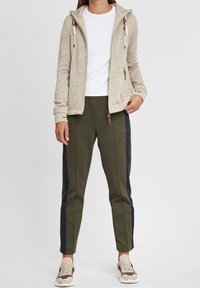 Giacca beige con cappuccio e coulisse, abbinata a pantaloni verde oliva con fondo stretto e strisce laterali blu navy, e sneakers beige.