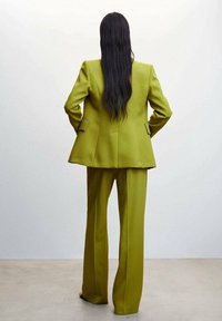 Persoon met lang donker haar, gekleed in een olijfgroen op maat gemaakt pak, staand met handen in de zij, met de rug naar voren tegen een effen achtergrond.
