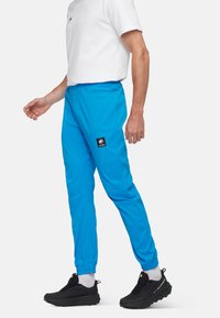 Un pantalon de sport bleu vif avec une taille à cordon, des jambes fuselées et un patch logo noir sur la cuisse gauche, associé à des chaussures noires et des chaussettes blanches.