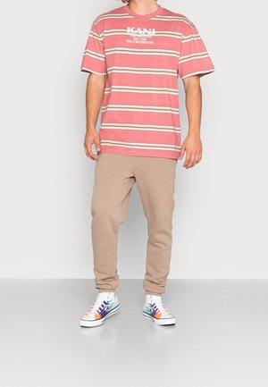 Man draagt een roze-wit gestreept KANI T-shirt, beige joggingbroek en witte sneakers met een blauw-oranje vlammenontwerp, staand op een grijze vloer.