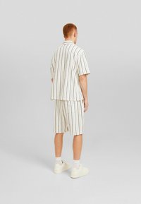 Man som bär en vit kortärmad skjorta och matchande shorts med vertikala bruna ränder, vita strumpor och vita sneakers, står vänd bort.