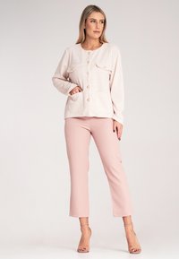 Jasnoróżowa kurtka zapinana na guziki z teksturowanej tkaniny, z dwiema kieszeniami na piersi i złotymi guzikami, w połączeniu z jasnoróżowymi spodniami typu cropped i cielistymi sandałami.