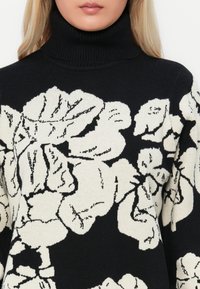 Melna augstākā kakla džemperis, kuram ir baltas ziedu raksti, adīta tekstūra un ribota apkakle. Dizains koncentrējas uz spēcīgu kontrastu un sarežģītiem detaļām.