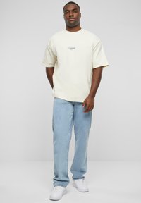 Cream oversized t-shirt with embroidered "Prohibited" logo, paired with light blue relaxed-fit jeans and white sneakers.