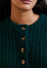 Donkergroene gebreide cardigan met een gestructureerd patroon, voorzien van vijf grote, ronde, bruine knopen aan de voorkant. Zacht, gezellig materiaal.