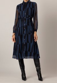 Button-up shirt dress in dark blue with geometric patterns, sheer long sleeves, and a matching fabric belt. Styled with black knee-high boots.