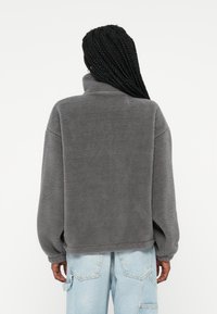 Pullover grigio in pile con colletto alto, maniche larghe e vestibilità rilassata. Il tessuto ha un aspetto morbido e testurizzato. Le cuciture visibili aggiungono struttura.