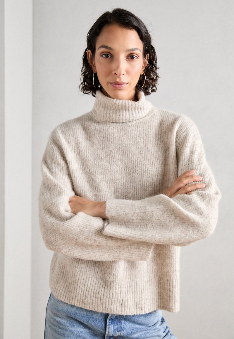 Kremfarget, ribbestrikket genser med høy krage, med avslappede ermer. Modellen står med armene i kors, iført blå jeans.