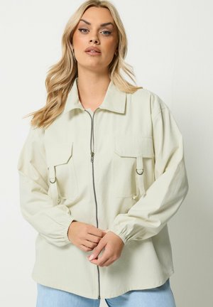 Lyshåret kvinde iført en off-white lynlåsjakke med store forlommer og ringe, poserende foran en ensfarvet hvid baggrund.