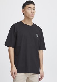 Black cotton t-shirt with short sleeves, loose fit, featuring a small green graphic design on the left chest area.