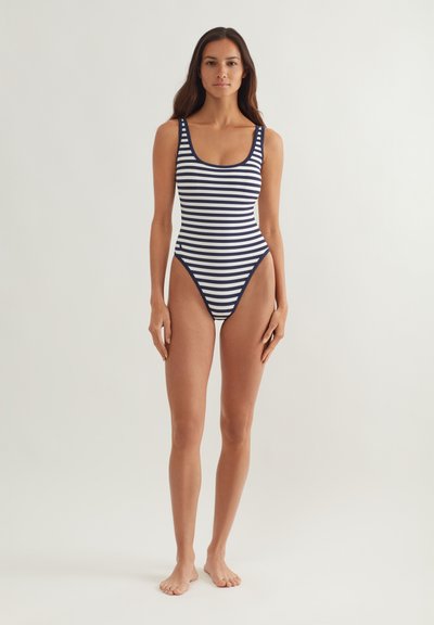 Γυναίκα όρθια ξυπόλητη, φορώντας ολόσωμο μαγιό με οριζόντιες ρίγες σε ναυτικό μπλε και άσπρο, μπροστά σε απλό ανοιχτό φόντο.