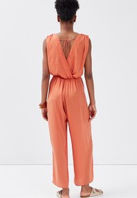 Combinaison rouge orangé sans manches, avec un dos profond en V, une taille élastique et un pantalon à jambes larges. Tissu lisse et léger.