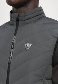 Muškarac nosi jaknu od šuškavog materijala u tamnosivoj boji s EA7 Emporio Armani logom i brendiranim patentnim zatvaračem.