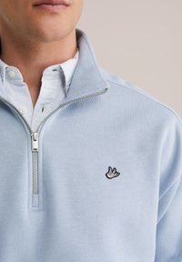 Šviesiai mėlynas užsegamas megztinis iš minkštos medžiagos su juodu siuvinėtu paukščio logotipu ir po juo baltu marškinių apykaklės dekoru.