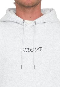 Hoodie gris clair à la texture douce, doté d'une capuche avec cordon de serrage et d'un logo "Volcom" brodé en noir sur la poitrine.