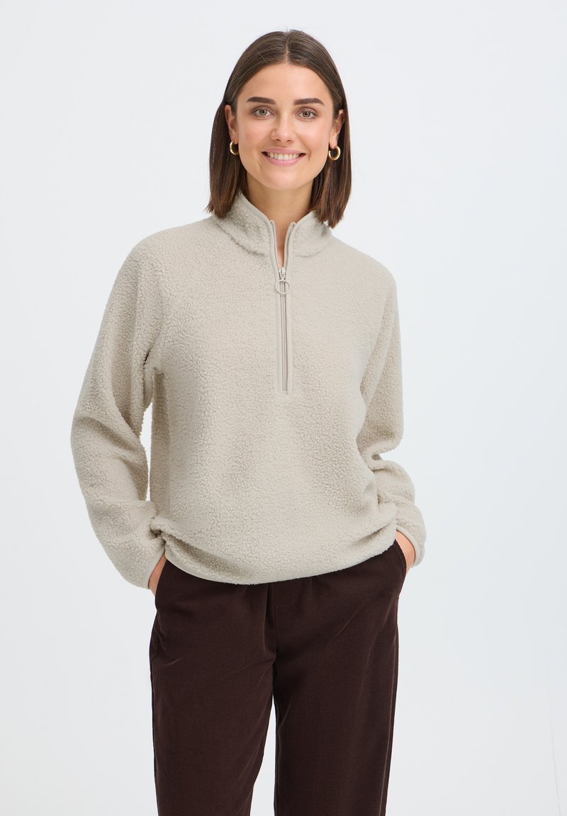 Femme aux cheveux courts et bruns portant des boucles d'oreilles créoles dorées, un pull polaire beige clair avec une fermeture éclair à moitié, et un pantalon marron foncé, les mains dans les poches.
