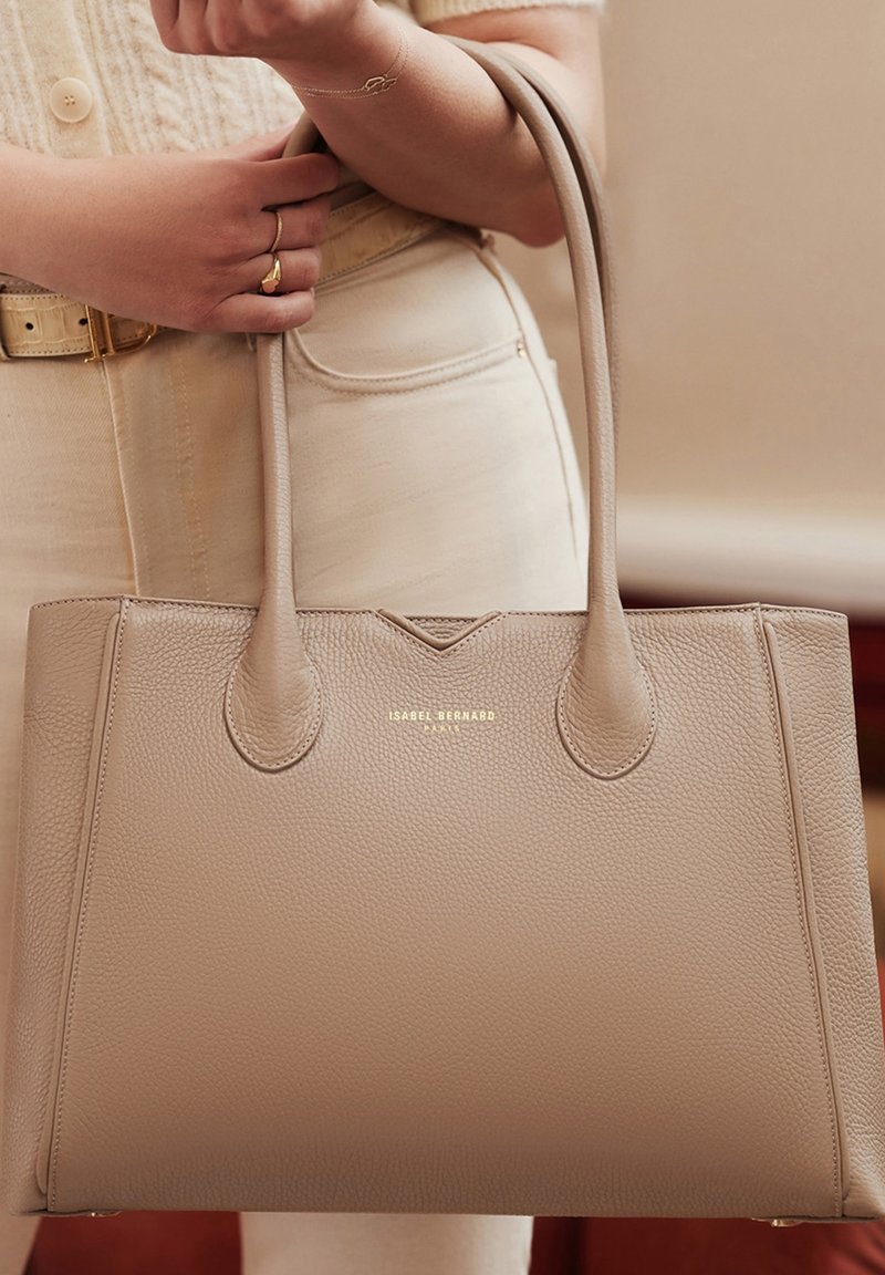 Beige leren tote bag met ronde handvatten, een gladde textuur en een reliëflogo aan de voorkant. De tas heeft een gestructureerde vorm en een minimalistisch ontwerp.