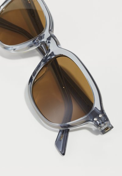 Sonnenbrille mit einem klaren grauen Kunststoffrahmen und braunen Gläsern. Besitzt eine markante Form und ein dezentes Logo-Detail am Bügel.