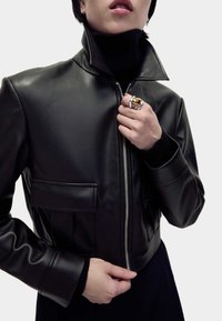 Schwarze Leder-Cropped-Jacke mit hohem Kragen, silbernem Reißverschluss und Fronttaschen. Das Modell präsentiert einen Ring mit goldfarbenem Akzent an der Hand.