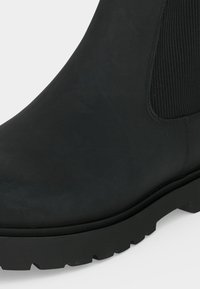 Gros plan de l'avant et du côté d'une botte Chelsea noire avec un panneau côtelé élastique et une semelle robuste sur fond blanc.