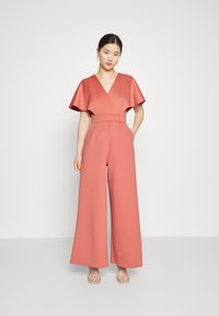 Ροζ φόρεμα με φαρδιά πόδια, V-λαιμόκοψη και φουσκωτά μανίκια. Φτιαγμένο από υφαντό ύφασμα; διαθέτει σφηκοφωλιά στη μέση και πλαϊνές τσέπες.