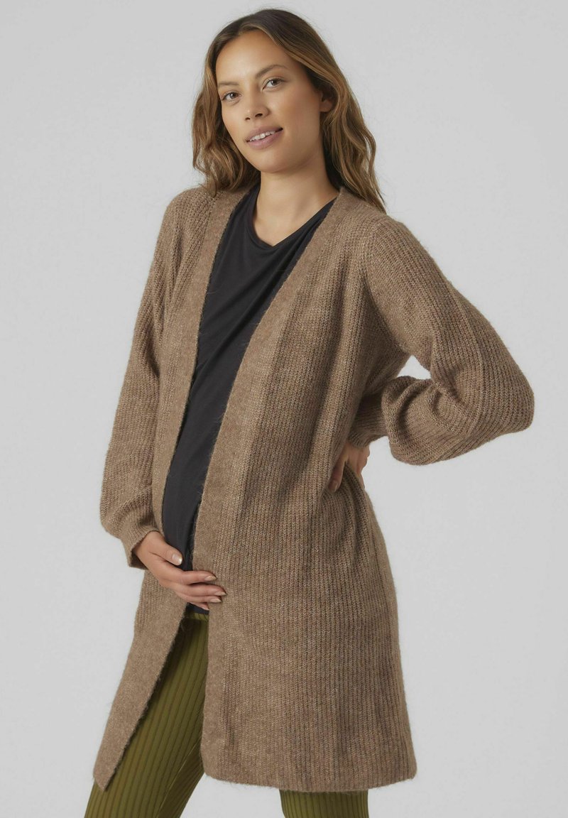 Cardigan marron à côtes avec manches longues et devant ouvert. Texture douce, longueur genoux, porté sur un haut noir et un pantalon vert à côtes.