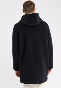 Homme portant un long manteau noir à capuche, vu de dos, se tenant devant un fond clair.